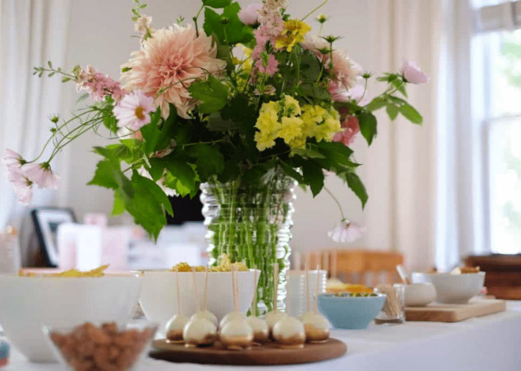 Large floral arrangement in the center of the table with gold and white cake pops in the foreground and other snacks in the beckground.