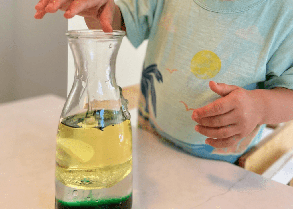 Child dropping an alka-seltzer tablet into a jar with water, green food coloring, and oil.