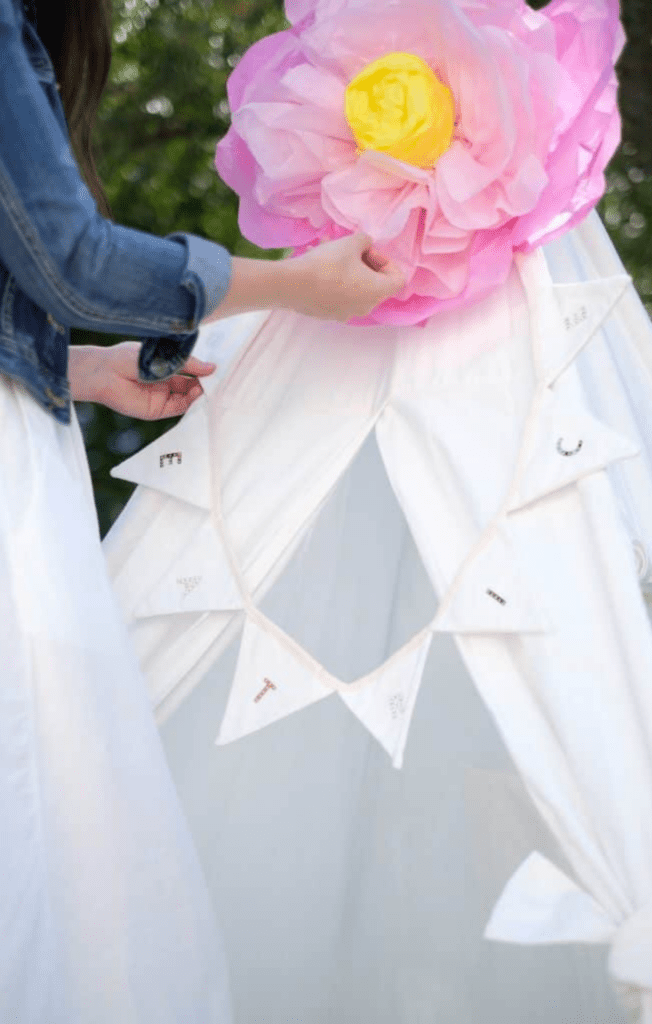 Woman adjusting a large tissue flower on the top of a white tipi-style tent.
