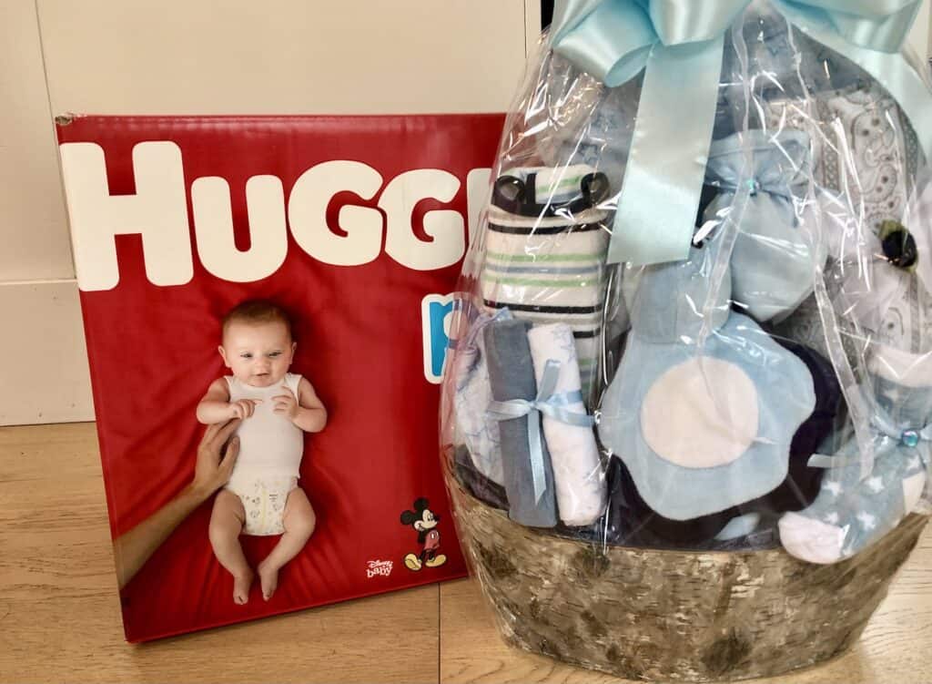 A box of huggies diapers next to a baby-themed gift basket with receiving blankets and a onsi, among other items.