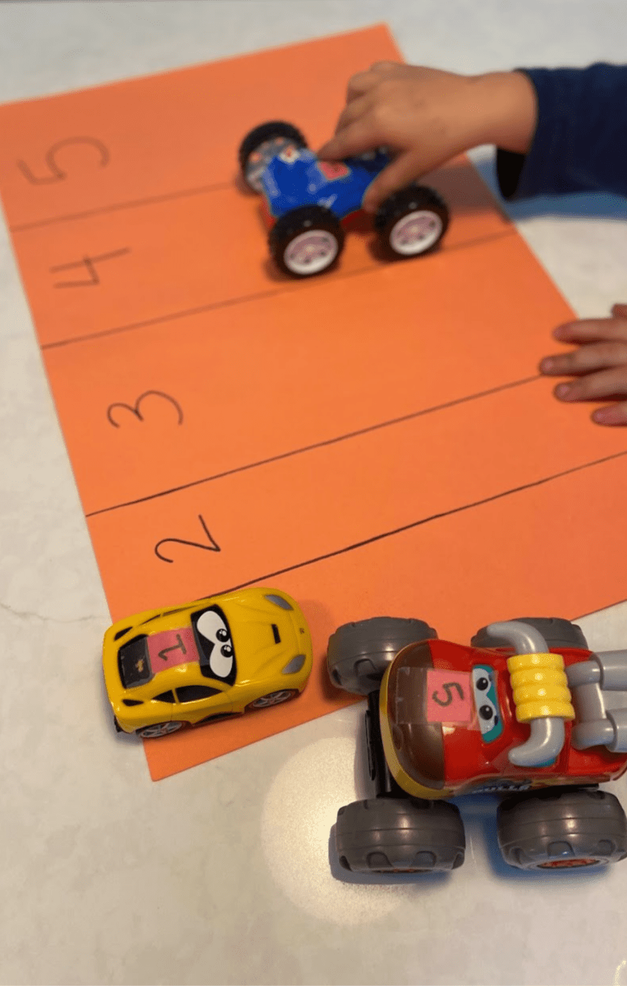 Orange piece of paper with numbers one through five written in black marker and a line separating each. Also pictures is a child organizing cars into the spaces that have corresponding numbers taped to the tops.