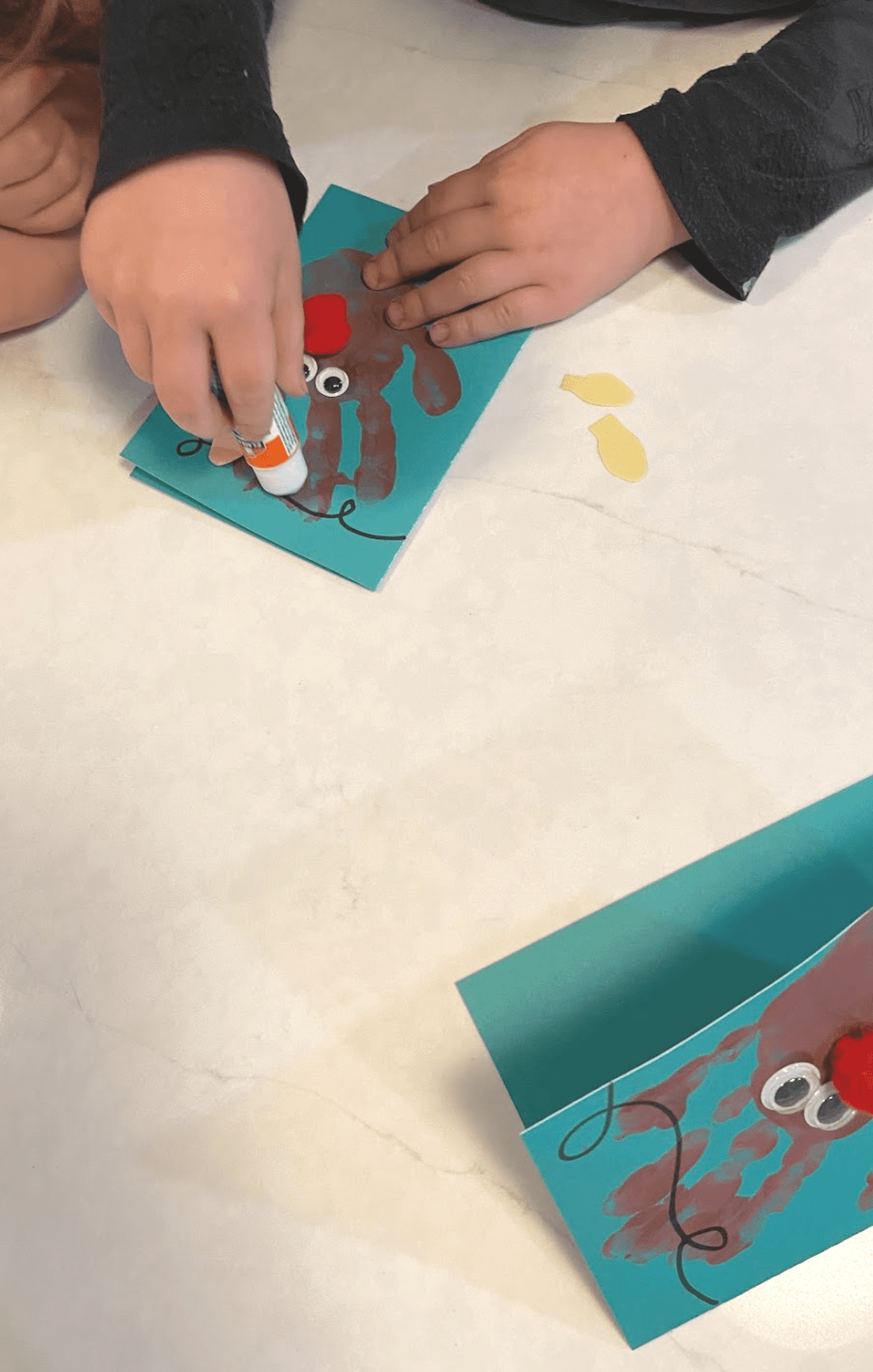 Handmade green cardstock Christmas cards with handprint reindeer. A child is gluing paper lights onto a hand-drawn string of lights.