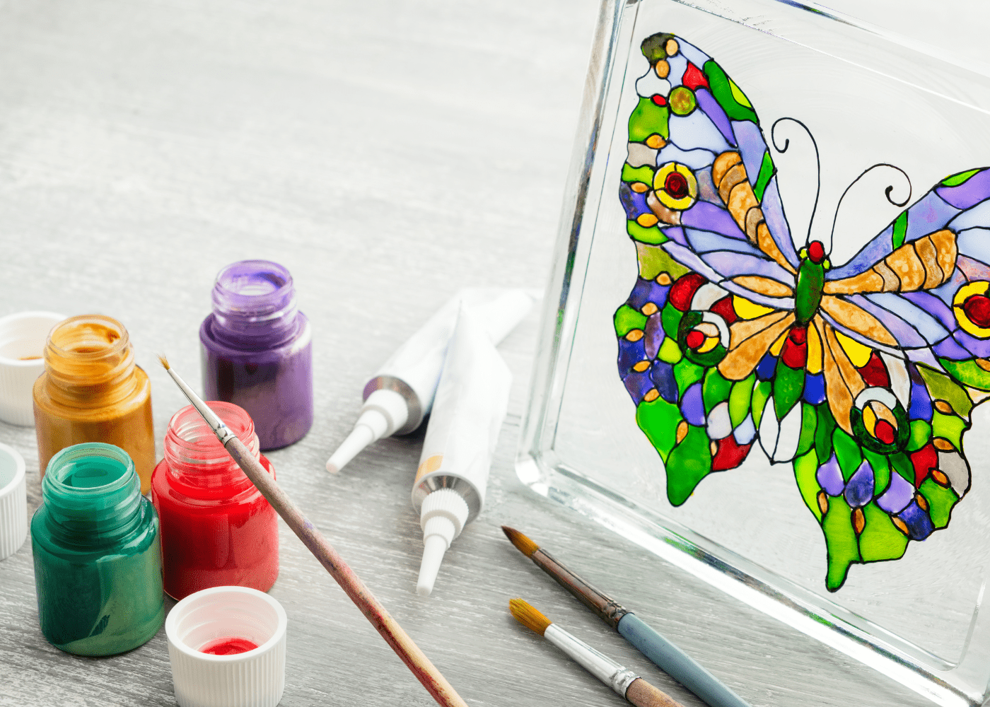 Photo of a stained glass butterfly on the right with the paints used, including gold, green, red, and purple.