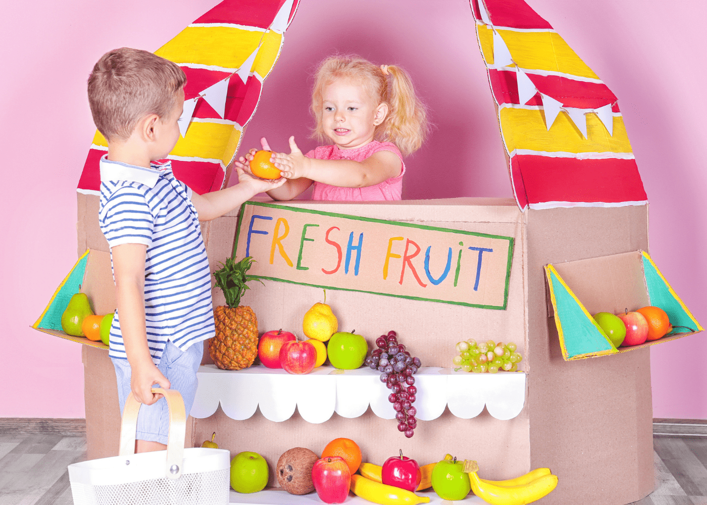 Two children in a carboard stand that says "fresh fruit. " they are pretending to buy and sell fake fruit.