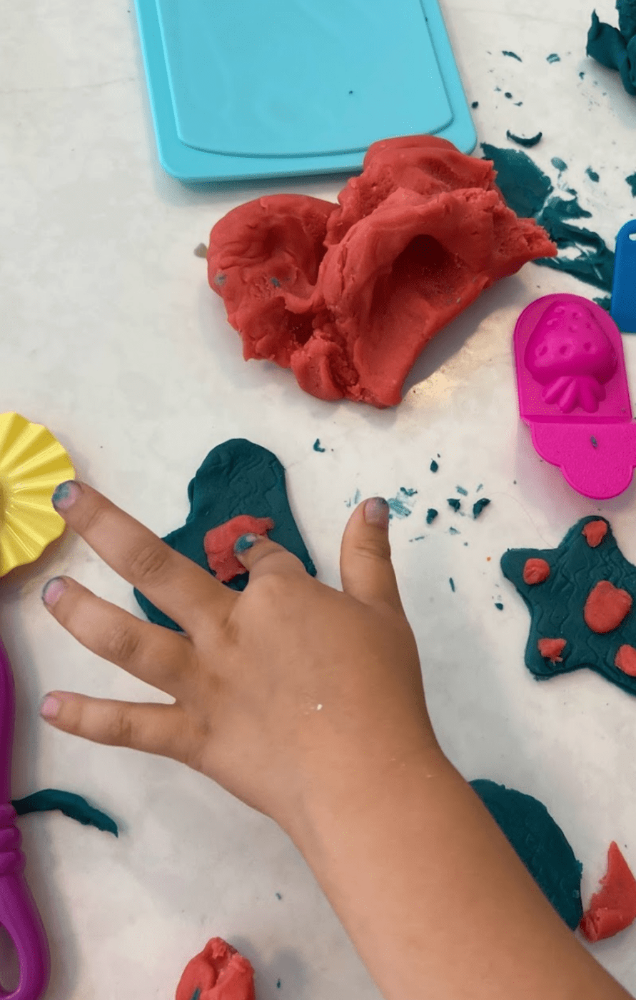A child' hand squishing a shape into a homemade playdough cutout of a heart. Other playdough and tools for manipulating it surround the child's hand.