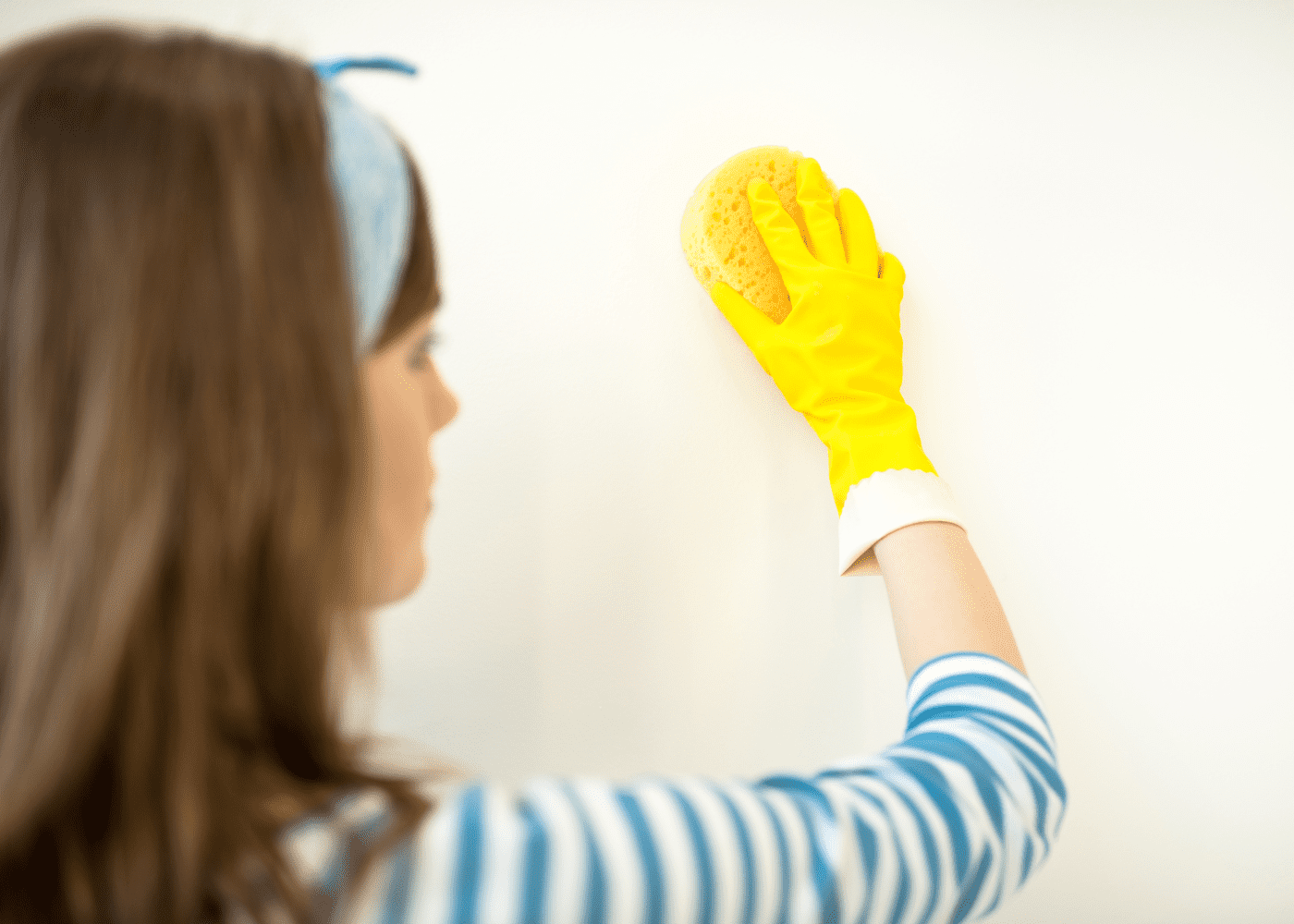 A woman cleaning a white wall with a sponge and yellow glove on in post how to clean white walls.