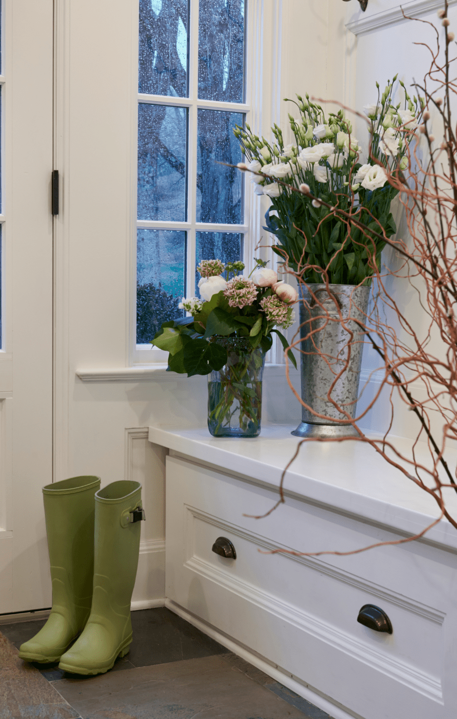 What are mudrooms? Image of two green boots beside a white bench with two floral arrangements.